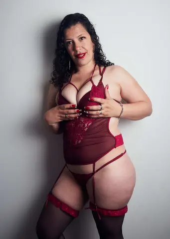Curvy woman in elegant red lace bodysuit and stockings posing against a white wall.