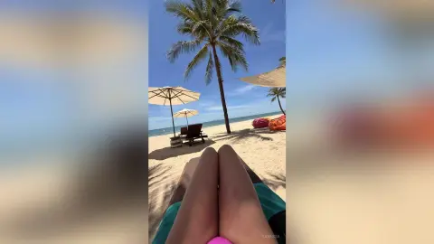 Relaxing view of sunbathing legs on a tropical beach with palm trees and white sand under a blue sky.