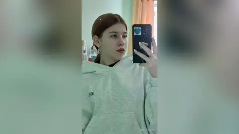 Young woman with reddish hair in a cozy grey hoodie taking a casual mirror selfie in a bright room.