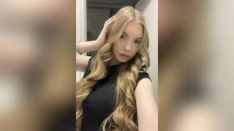ViolaFausel poses with her long blonde wavy hair, wearing a black t-shirt against a plain white wall.