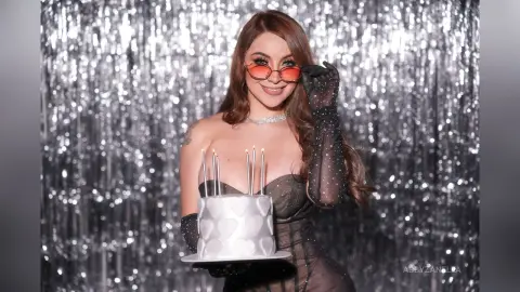 Glamorous woman in black sheer outfit holding a white cake against a sparkling silver tinsel backdrop.