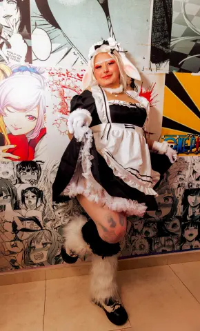 Cosplayer in a black and white maid outfit with bunny ears posing in front of anime manga wall art.