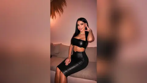 BiankaLeoness poses in a black leather crop top and tight leather skirt while sitting on a couch.