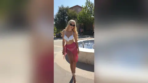 Blonde woman in a white bikini top and pink sarong posing by a sunny outdoor fountain.