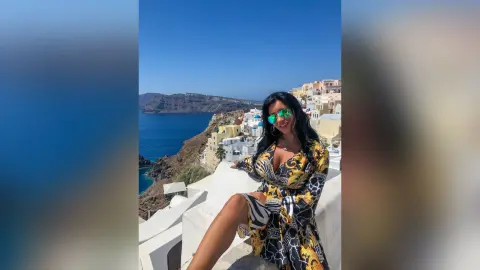 MissyJolie poses on a white balcony overlooking the blue sea, wearing a black and yellow patterned wrap dress and green sunglasses.