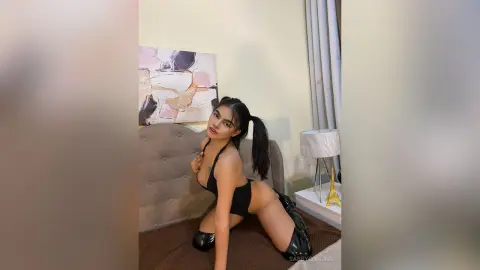 Young woman in a black bodysuit posing on a bed in a modern bedroom setting.