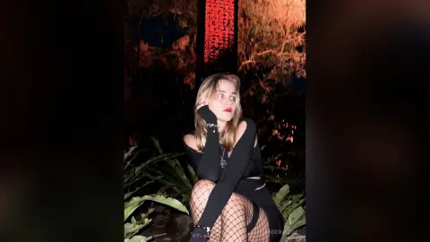 Young woman in a black crop top and fishnet tights posing moody against a dark night garden background.