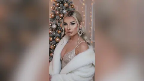 Glamorous blonde woman in a white faux fur coat posing by a decorated Christmas tree in a festive setting.