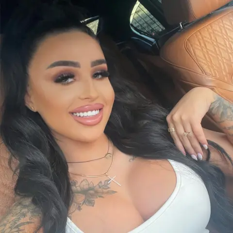 Smiling woman with long dark hair and tattoos wearing a white top in a luxury car interior.