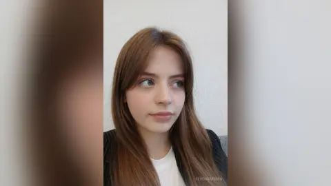 Young woman with long brown hair wearing a black blazer and white shirt in a minimalist indoor setting.