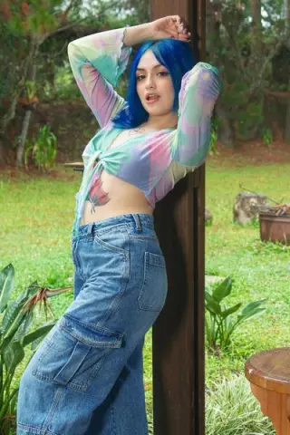 Young woman with blue hair in a colorful tie-dye crop top and denim jeans posing outdoors in a sunny garden.