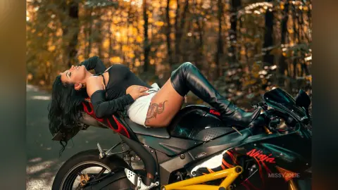 MissyJolie reclines nakedly across a black motorcycle, wearing black fingerless gloves and long black boots against a sunlit forest background.