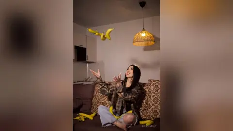 JuliDeming sits on a patterned couch in a dark room, laughing as she tosses yellow bananas into the air.