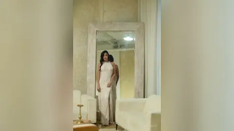 Elegant woman in a white sleeveless gown standing before a large mirror in a bright, minimalist modern living room.