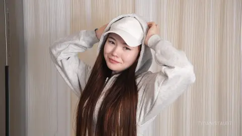 Smiling young woman in a cozy grey hoodie and white baseball cap posing against neutral striped curtains.