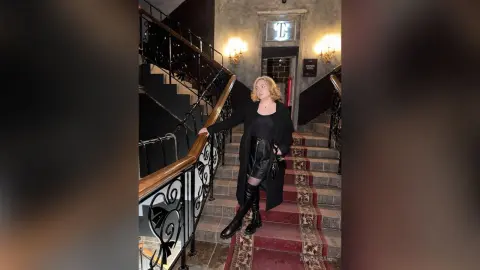 AnnyFraid stands on a carpeted staircase wearing a black long coat, black top, black leather skirt, and tall black boots.