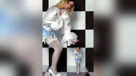 Blonde woman in a white puff-sleeve blouse and blue lace skirt posing against a black and white checkered backdrop.