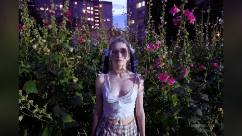 Young woman in a white lace camisole standing in a dreamy garden of pink hollyhock flowers at twilight.