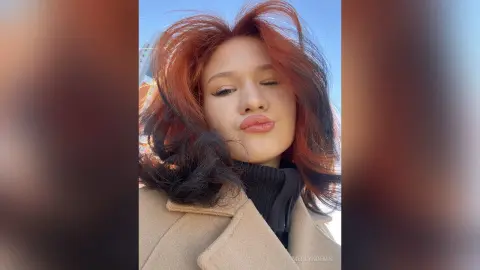 Young woman with reddish-brown hair wearing a black turtleneck and tan coat outdoors in soft sunlight.