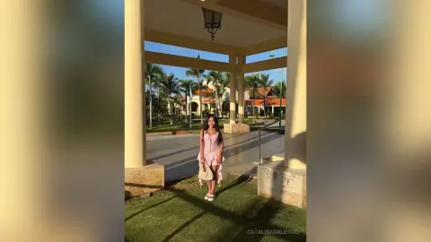 Woman in a pink summer dress standing under a sunny outdoor patio with palm trees in the background.