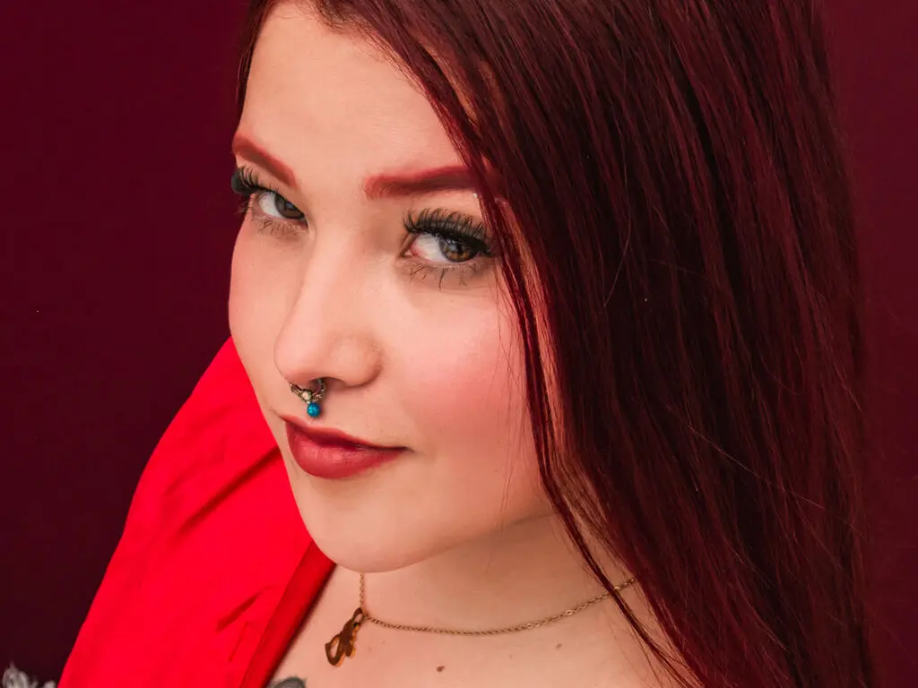 Close-up portrait of a woman with red hair and nose ring wearing a red outfit against a dark background.