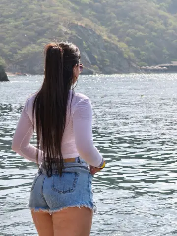 Woman in white long-sleeve shirt and denim shorts looking at a sparkling lake near lush green mountains.