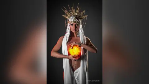 LeslyAnderson wears a gold spiked headpiece and white draping, holding a glowing orange orb against her bare chest and stomach.