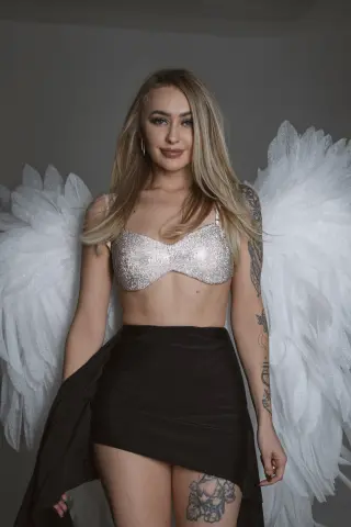 Blonde woman in a silver sequin bikini top and black skirt wearing large white angel wings in a studio setting.