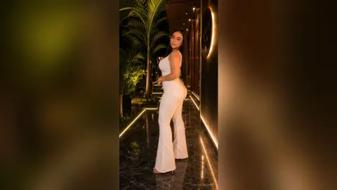 NatalyBloom poses in a tight white jumpsuit at night, standing on a dark patio next to palm trees.