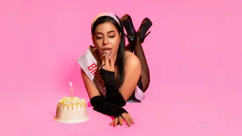 Young woman in white birthday sash lying on pink background with a birthday cake in a playful mood.
