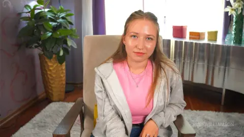 RubeWilliamson wearing a pink t-shirt and a light grey zip-up hoodie while sitting in a chair in a room with a plant and a silver dresser.