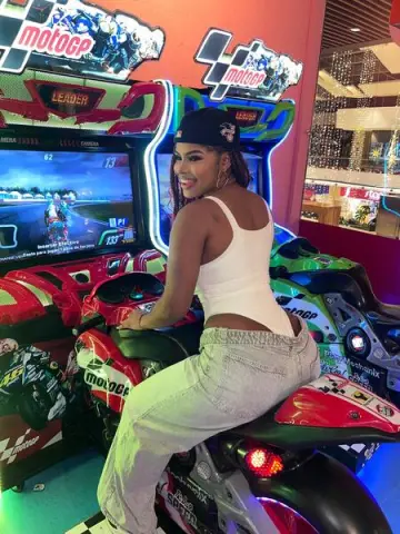 Smiling woman in white tank top and jeans playing a MotoGP racing arcade game in a bright gaming lounge.