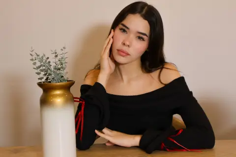 Young woman in a black off-the-shoulder top sitting by a gold vase in a minimalist indoor setting.