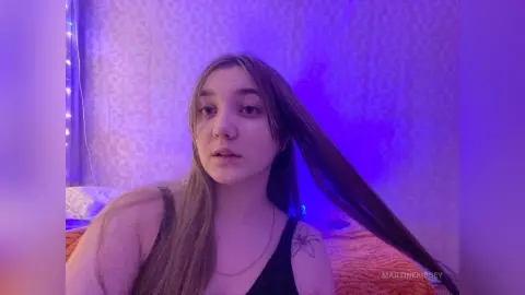 MartineKibbey looks at the camera with long brown hair and a black tank top, lit by blue light in a bedroom setting.