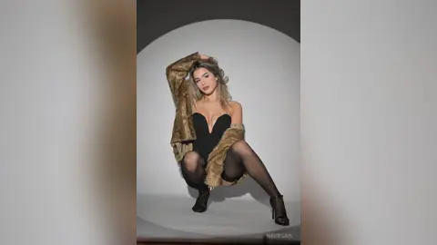 Glamorous woman in a black bodysuit and faux fur coat posing in a studio setting.