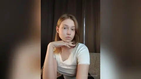 EvelinaMoorina sits on a couch wearing a white lace top and looking into the camera.