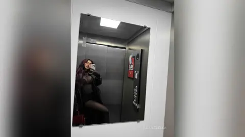 NicoleBella poses for a mirror selfie in an elevator wearing a black sheer bodysuit and black tights.