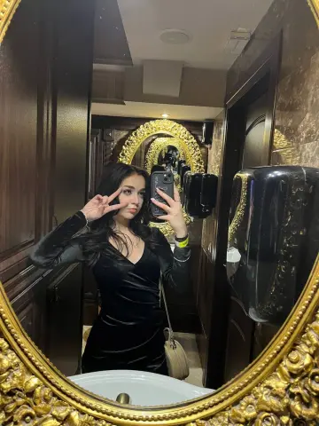 Young woman in a black velvet dress taking a mirror selfie in a luxury bathroom with a gold ornate mirror.