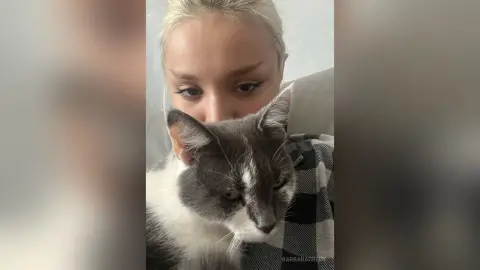 BarbaraGreen poses closely with a grey and white cat, showing her blonde hair and eyes while wearing a black and white plaid shirt.
