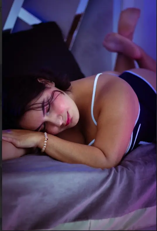 Curvy woman in black and white lingerie relaxing on a bed in soft, moody purple lighting.