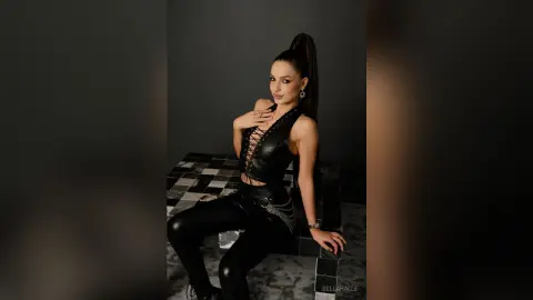 Stylish woman in black leather outfit posing confidently on a checkered platform in a moody studio setting.
