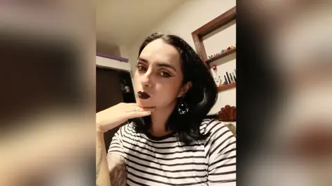 Young woman with dark hair and bold lipstick wearing a striped shirt in a cozy indoor setting.