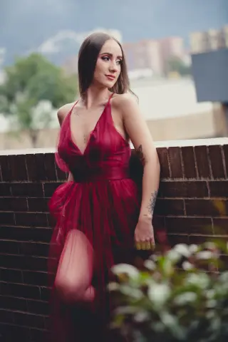 Elegant woman in a flowing red evening gown posing by a brick wall in a soft outdoor setting.
