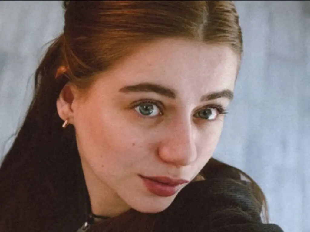 Close-up portrait of a young woman with brown hair and blue eyes wearing a black sweater in soft natural light.