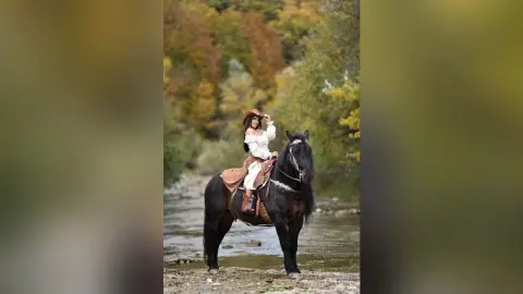 Woman in white boho blouse riding a black horse through a scenic autumn forest river landscape.