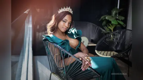 Elegant woman in a teal ruffled gown and silver crown sitting in a modern lounge setting.