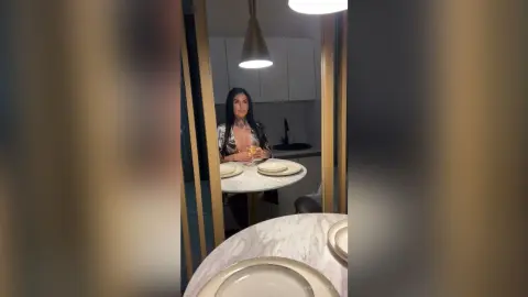 Elegant woman in a patterned blouse sitting at a marble dining table in a modern, dimly lit kitchen.