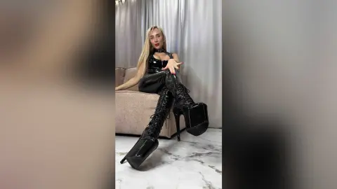 Blonde woman in edgy black leather outfit and high platform boots sitting on a sofa in a bright room.