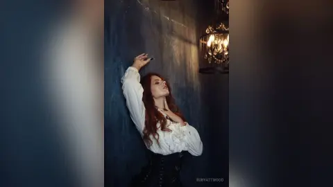 RubyAttwood poses against a dark wall wearing a white blouse and a black corset, running her hand through her long red hair.