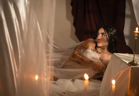 Tattooed woman in white flowing fabric lounging in a moody, candlelit room with draped curtains.